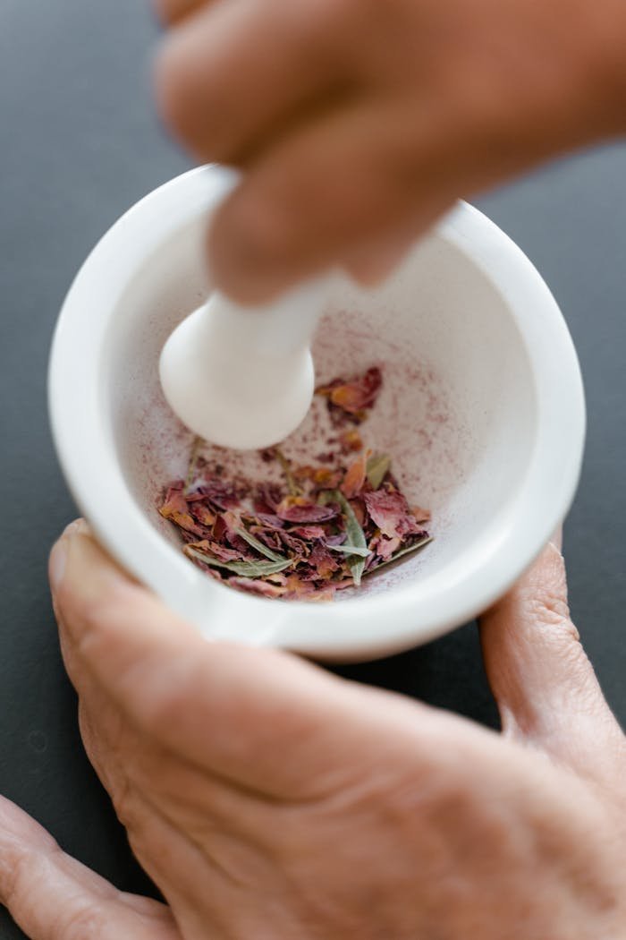 A hand using a mortar and pestle to crush dried flower petals for body care products.